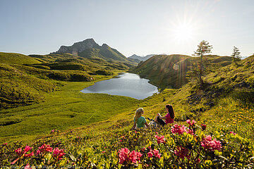 Frauen sitzen inmitten von Almrosen und schauen auf einen Bergsee