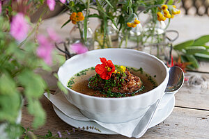 Eine köstliche Schüssel Kaspressknödelsuppe, garniert mit frischen Kräutern und bunten essbaren Blumen, steht auf einem rustikalen Holztisch, umgeben von zarten Blumenarrangements, die eine einladende Atmosphäre schaffen.