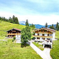 Appartement mieten in Fieberbrunn,Tirol Zwei charmante Alpenhäuser stehen in einer malerischen Landschaft, umgeben von saftigem Grün und blühenden Bäumen. Der strahlend blaue Himmel sorgt für eine friedliche Atmosphäre, während die Berge im Hintergrund eine beeindruckende Kulisse bieten.