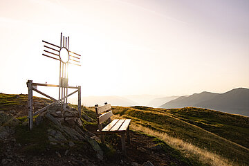Ein Gipfelkreuz mit Bankerl in goldenem Sonnenlicht