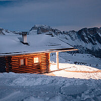 Beleuchtete Almhütte im Winter bei Nacht, im Hintergrund verschneite Bergwelt