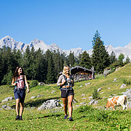 Zwei Frauen beim Wandern, im Hintergrund sieht man eine urige Almhütte